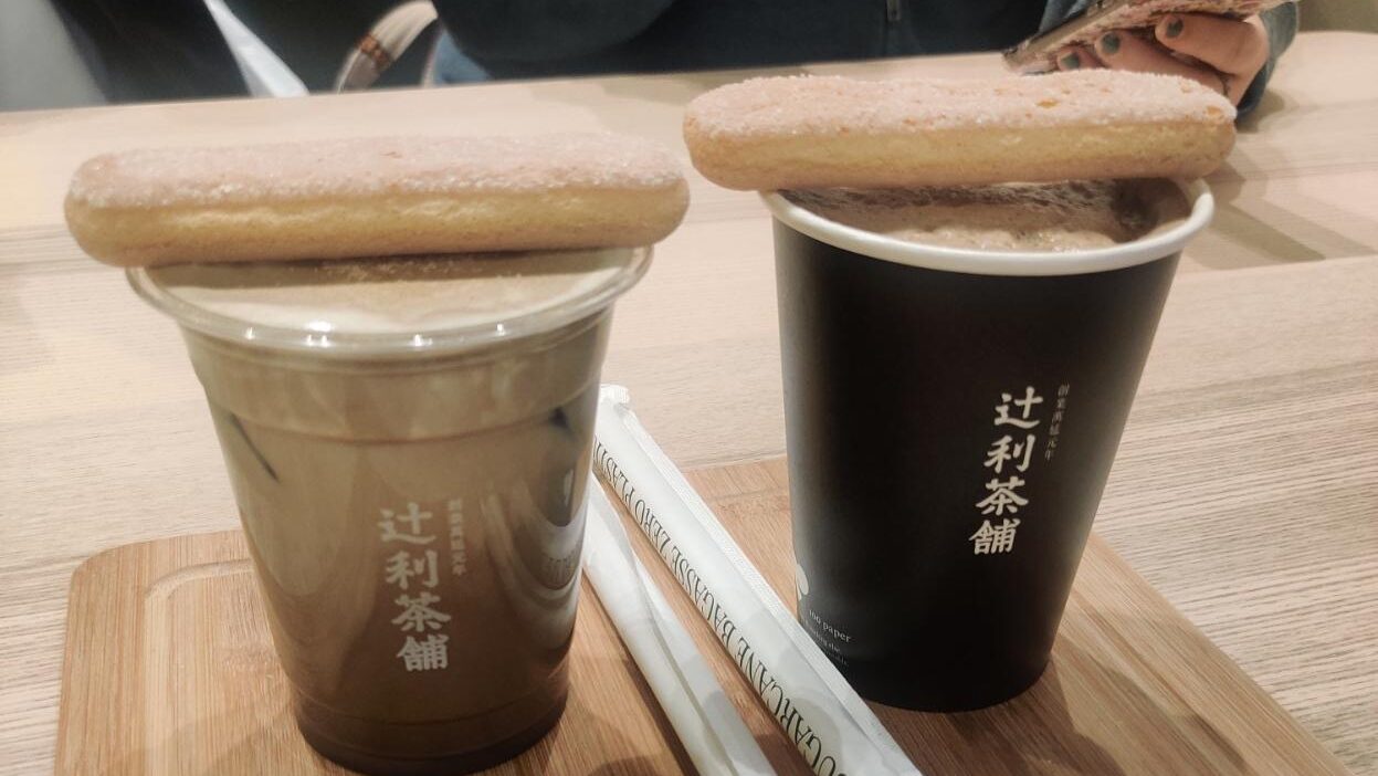 two cups of tiramisu hojicha, the one of the left is cold in a plastic cup, the one on the right is hot in a paper cup, and there is a garibaldi biscuit lying on top of each cup