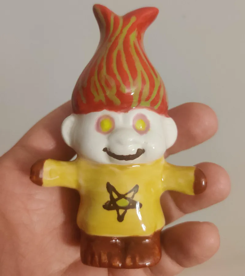 a small goblin figurine hand painted to look like. a demon: the hair is red with green streaks, the eyes are yellow with a purple circle around them, the hands and feet are a brown red and the jumper is yellow with the same brown red colour depicting a pentagram